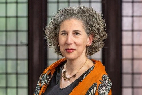 A woman with curly grey hair and a slight smile wearing a necklace and orange and black outfit stands in front of windows with square muntins. 