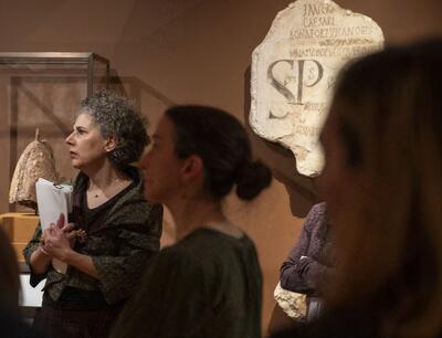 A group of people in a museum looking to the left. A woman in standing prominently in the light. Behind them is a piece of Roman graffiti with the letters SP visible.