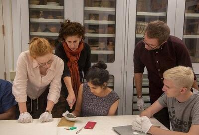 A group of students with a professor examine items in a museum research area.